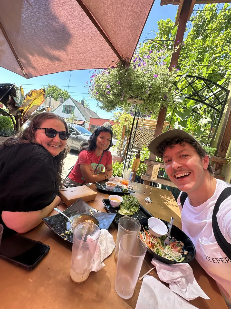 Three people smiling at an outdoor dining table in a lush garden setting under a red umbrella in Portland.