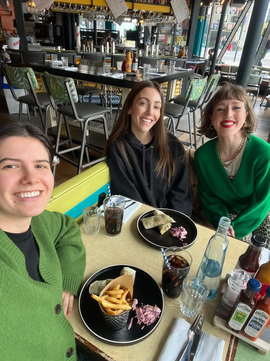 Three smiling women at a London bar restaurant during a Creative Lunch Club meetup, with food and drinks on the table.