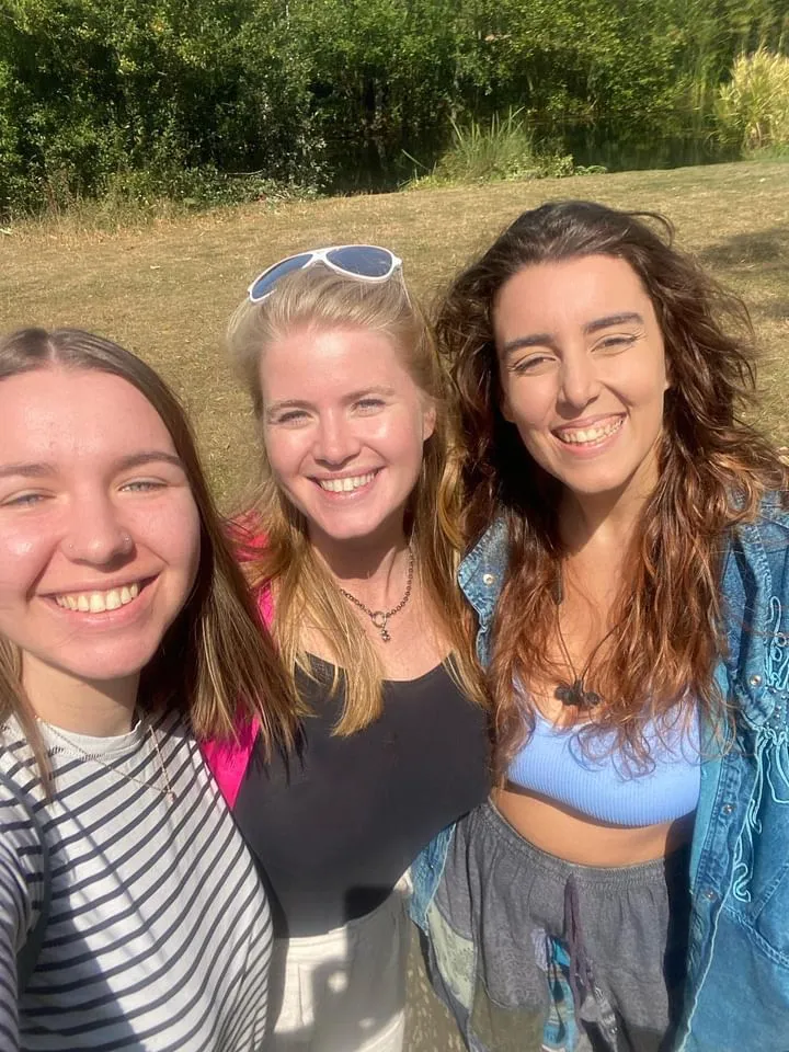 Three smiling young women posing outdoors on a sunny day with green trees in the background at a Creative Lunch Club meetup.