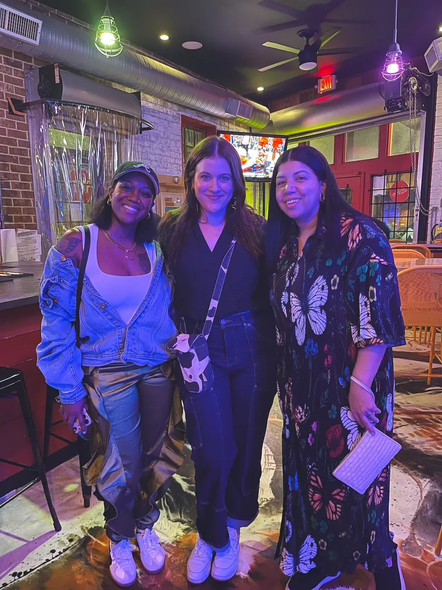 Three women smiling at a Creative Lunch Club meetup in Atlanta, in a vibrant industrial venue with neon lighting.