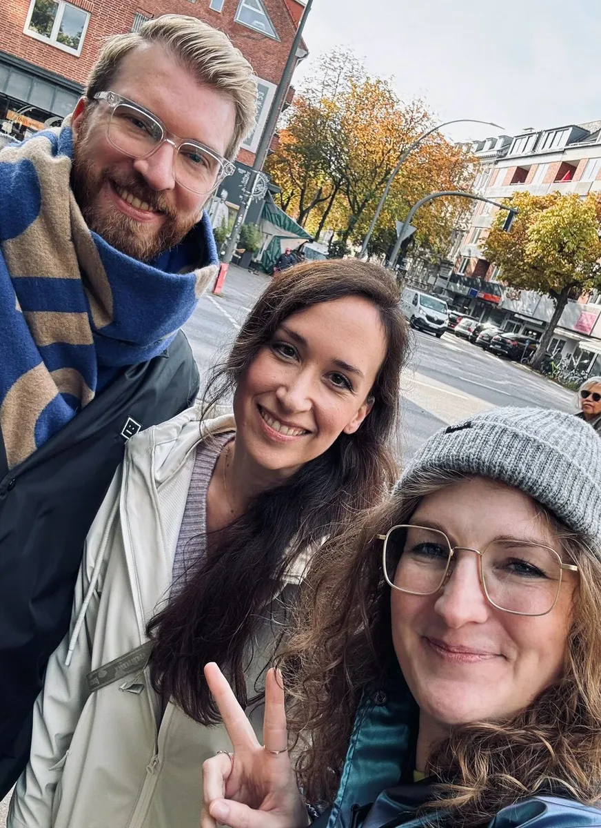 Three people smiling at a Creative Lunch Club meetup in Hamburg, with autumn trees and brick buildings in the background.