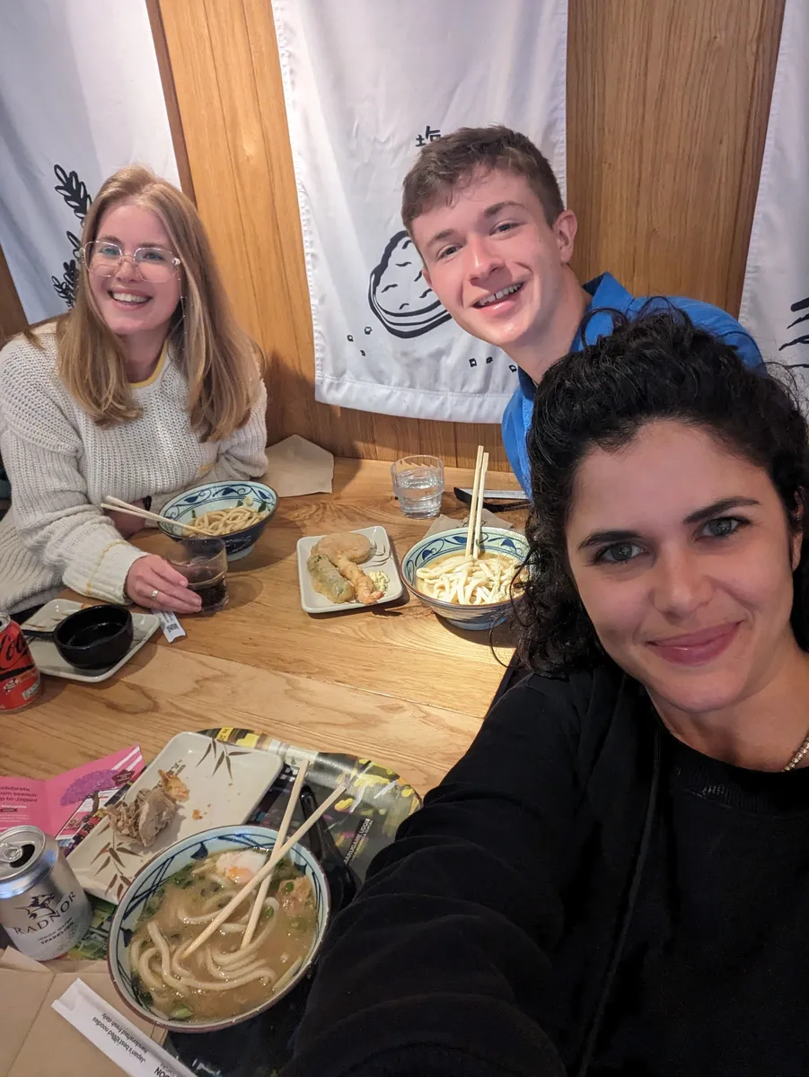 Three people enjoying noodles at a Creative Lunch Club meetup in London, smiling at a wooden table.