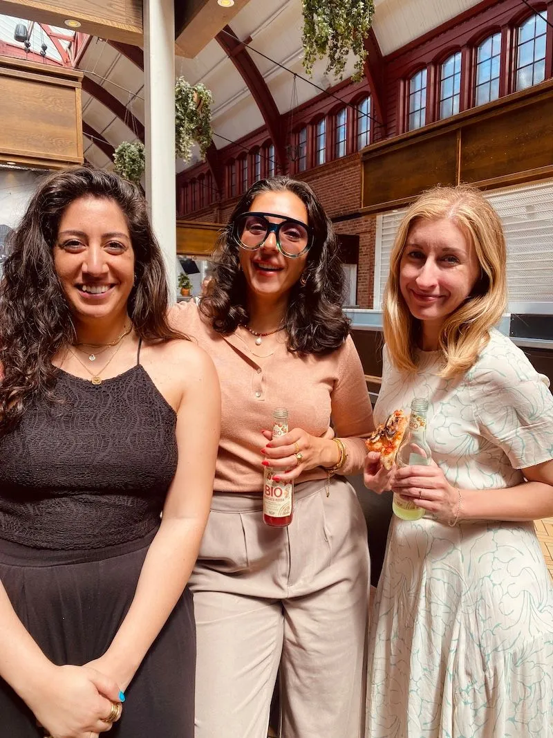 Three women smiling at a Creative Lunch Club meetup in Malmö, standing in a modern brick venue with high ceilings and hanging plants.