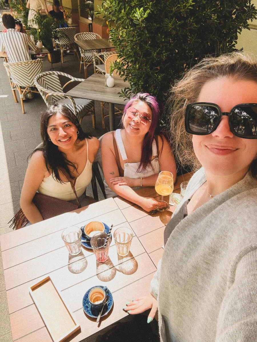 Three women smiling at a sunny outdoor café terrace in Budapest with drinks and pastries on the table.
