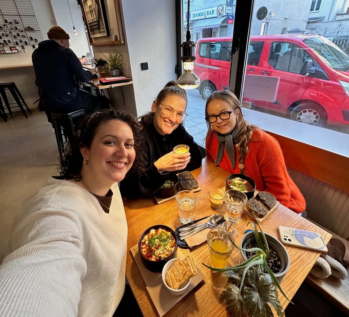 Three smiling women enjoying lunch together at a Munich café with red vehicles visible through the window behind them.