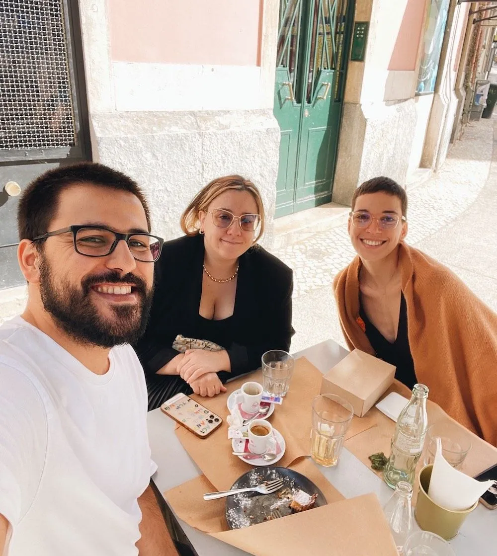 Three smiling people enjoying coffee and pastries at an outdoor café in Lisbon during a Creative Lunch Club meetup.