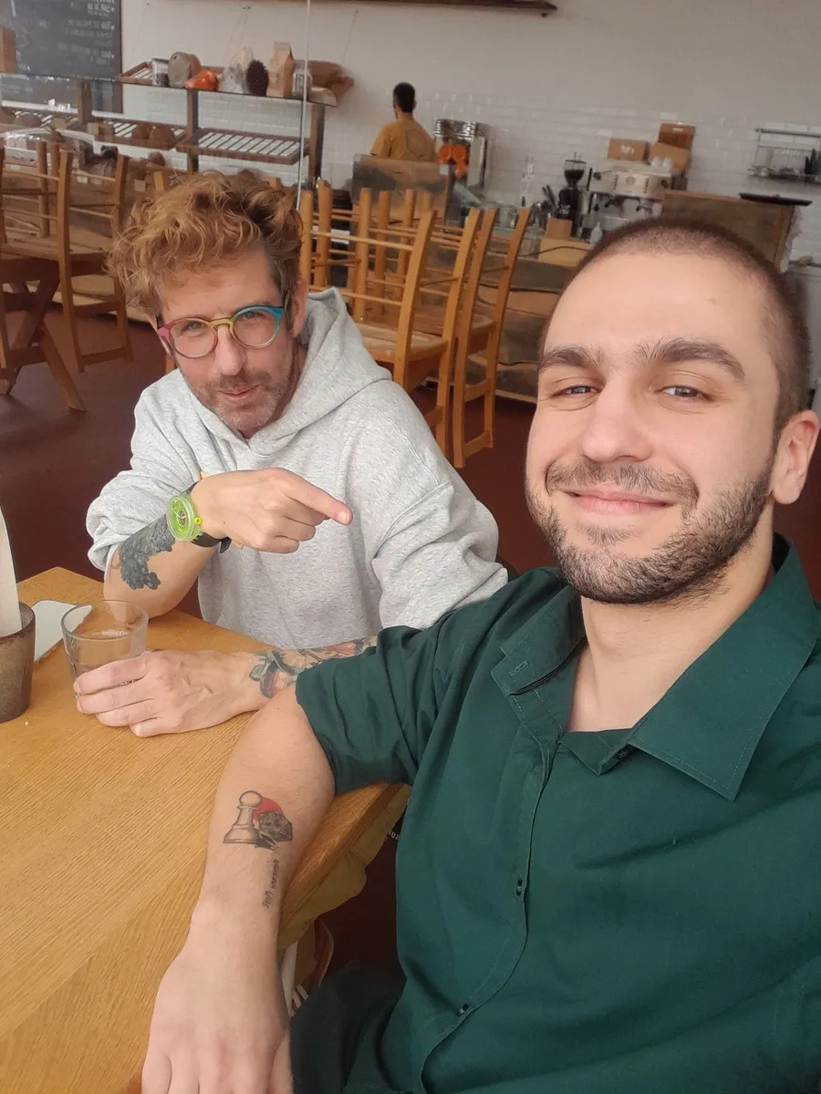 Two men smiling at a Creative Lunch Club meetup in Porto, seated at a wooden table in a casual dining space with chairs in background.