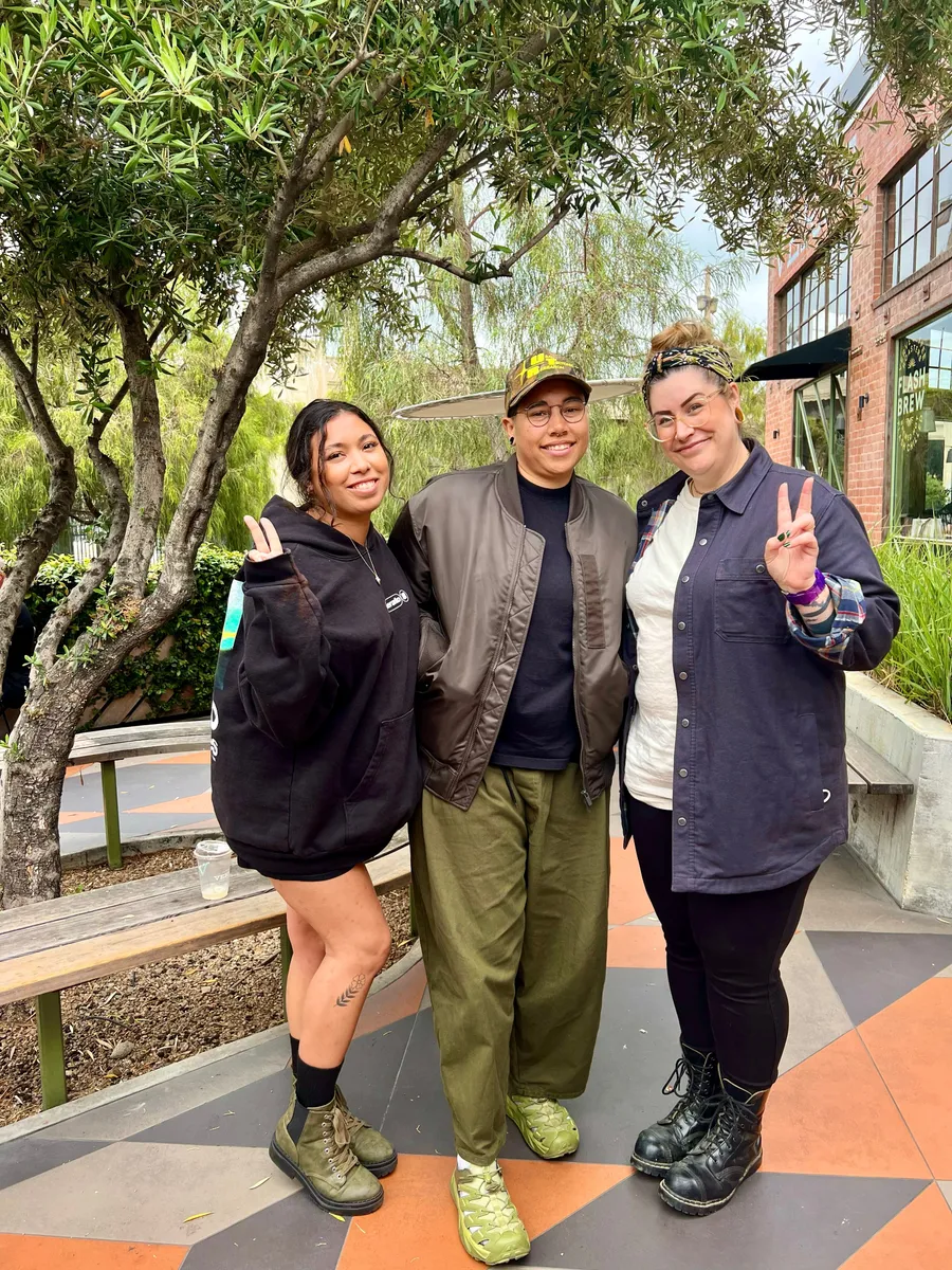 Three people smiling at a Creative Lunch Club meetup in Los Angeles, standing under trees with brick buildings in background.