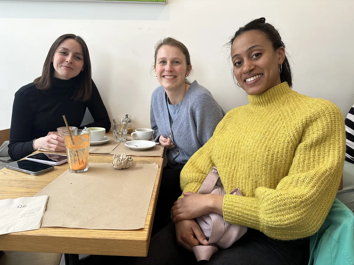 Three smiling women at a wooden table during a Creative Lunch Club meetup in Brussels, with beverages and notebooks.
