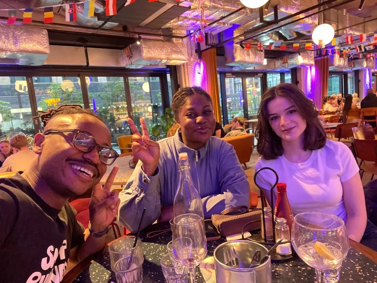 Three people smiling at a dining table during a Creative Lunch Club meetup in Manchester, purple-lit rooftop venue with city views.