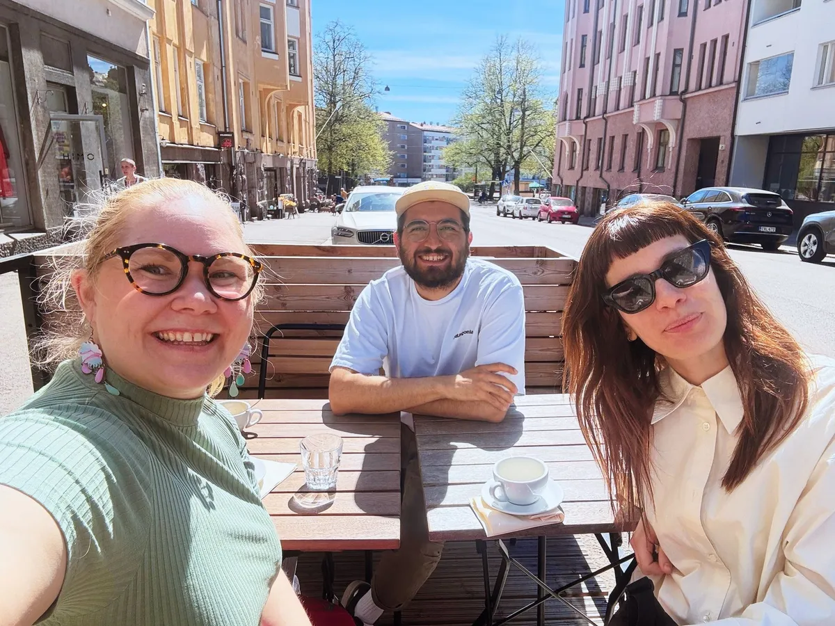 Three people smiling at outdoor café table in sunny Helsinki street with colorful buildings in background.
