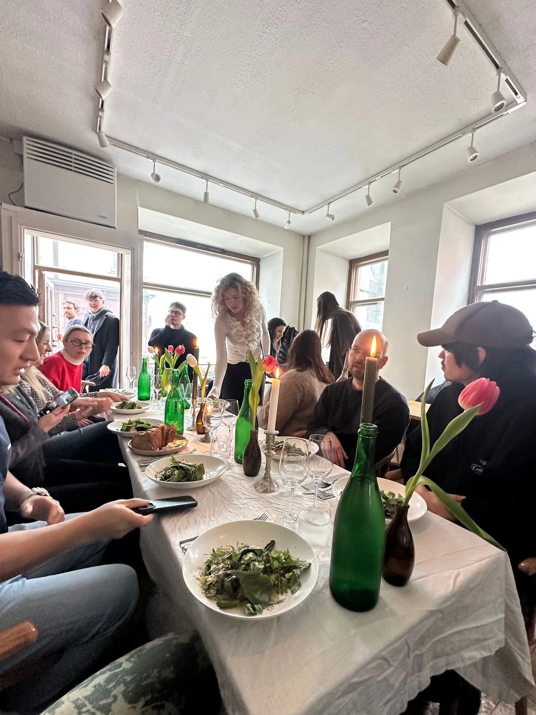 Creative professionals networking at a Helsinki lunch meetup, seated at a long white-clothed table with salads, green bottles, and pink tulips.