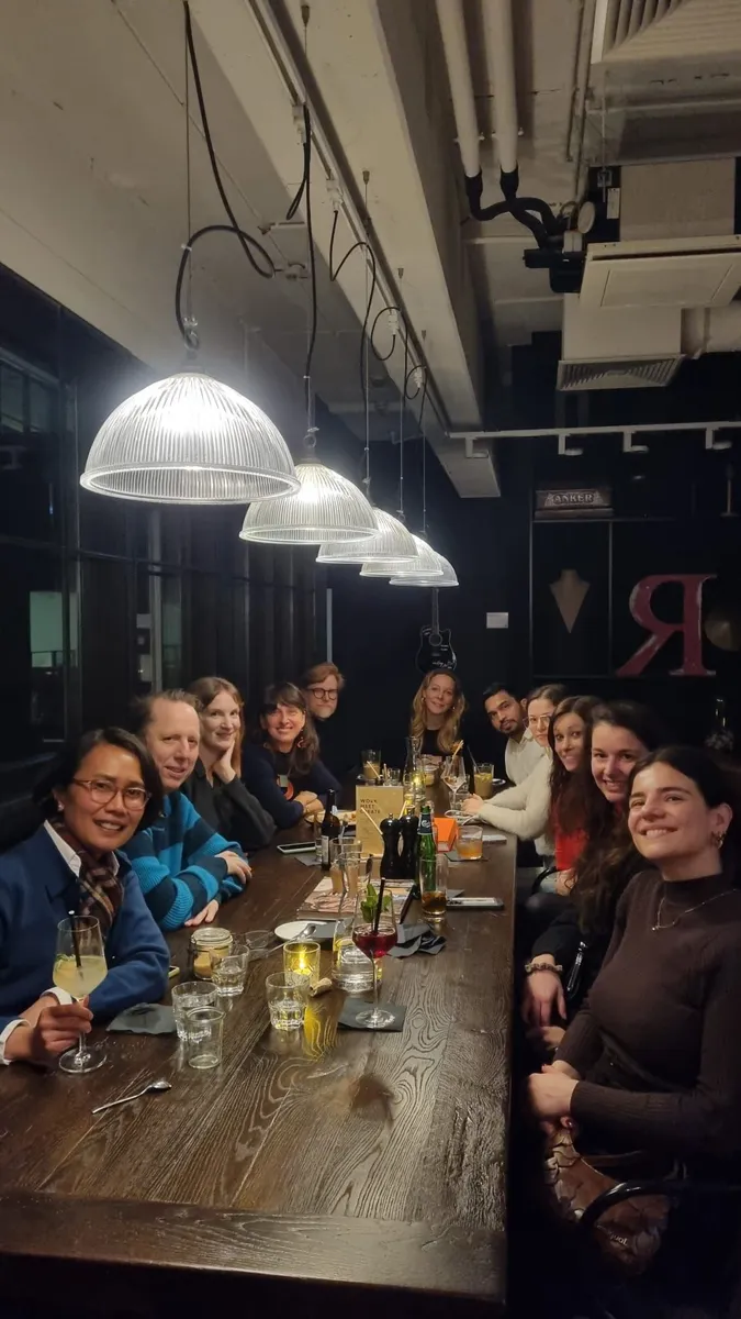 A group of creative professionals gathered around a long wooden table at a Creative Lunch Club meetup in Düsseldorf, enjoying drinks and conversation under modern pendant lighting.