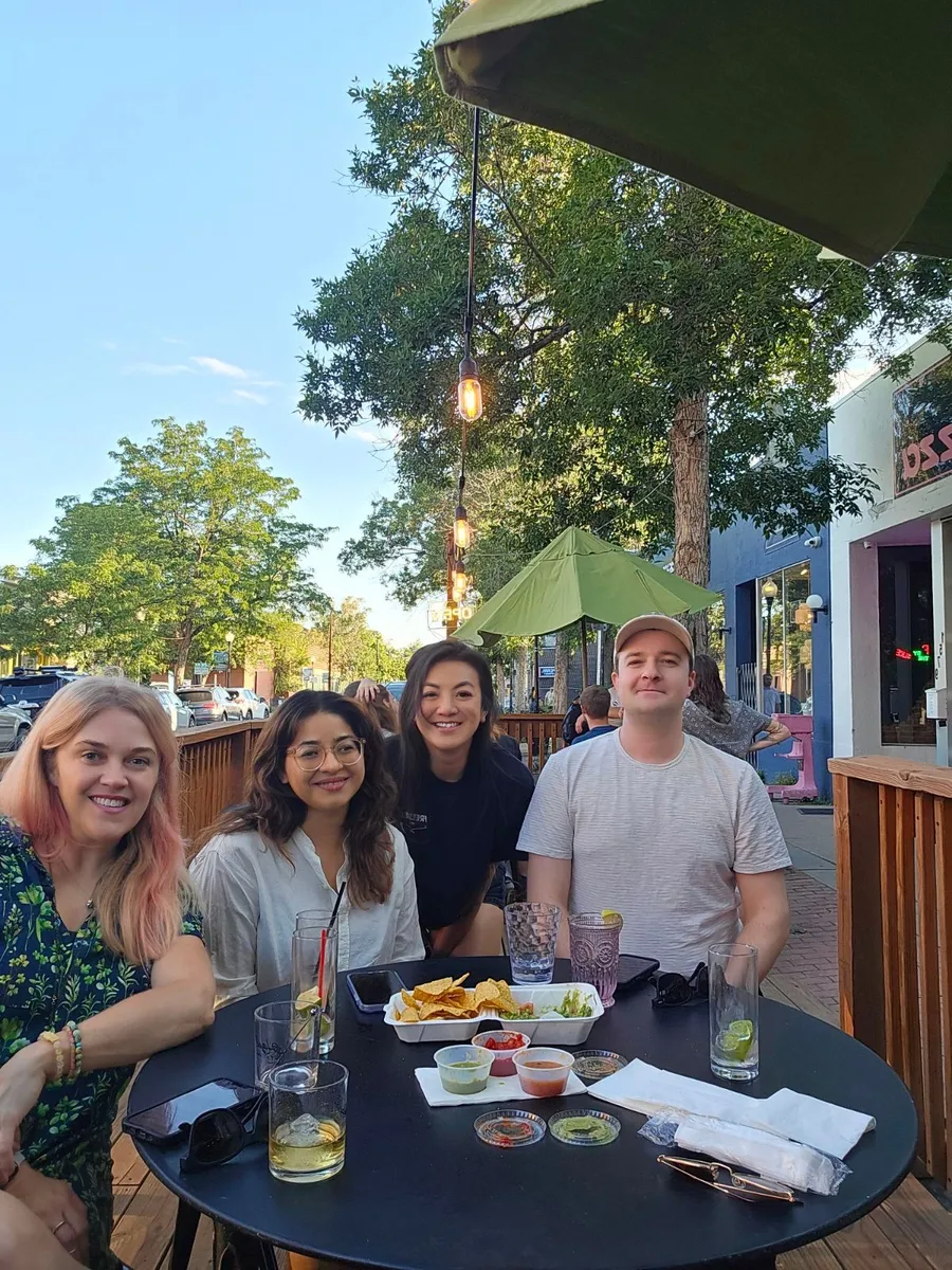 Four young professionals smiling at outdoor patio table during Creative Lunch Club meetup in Denver, with trees and storefronts visible.