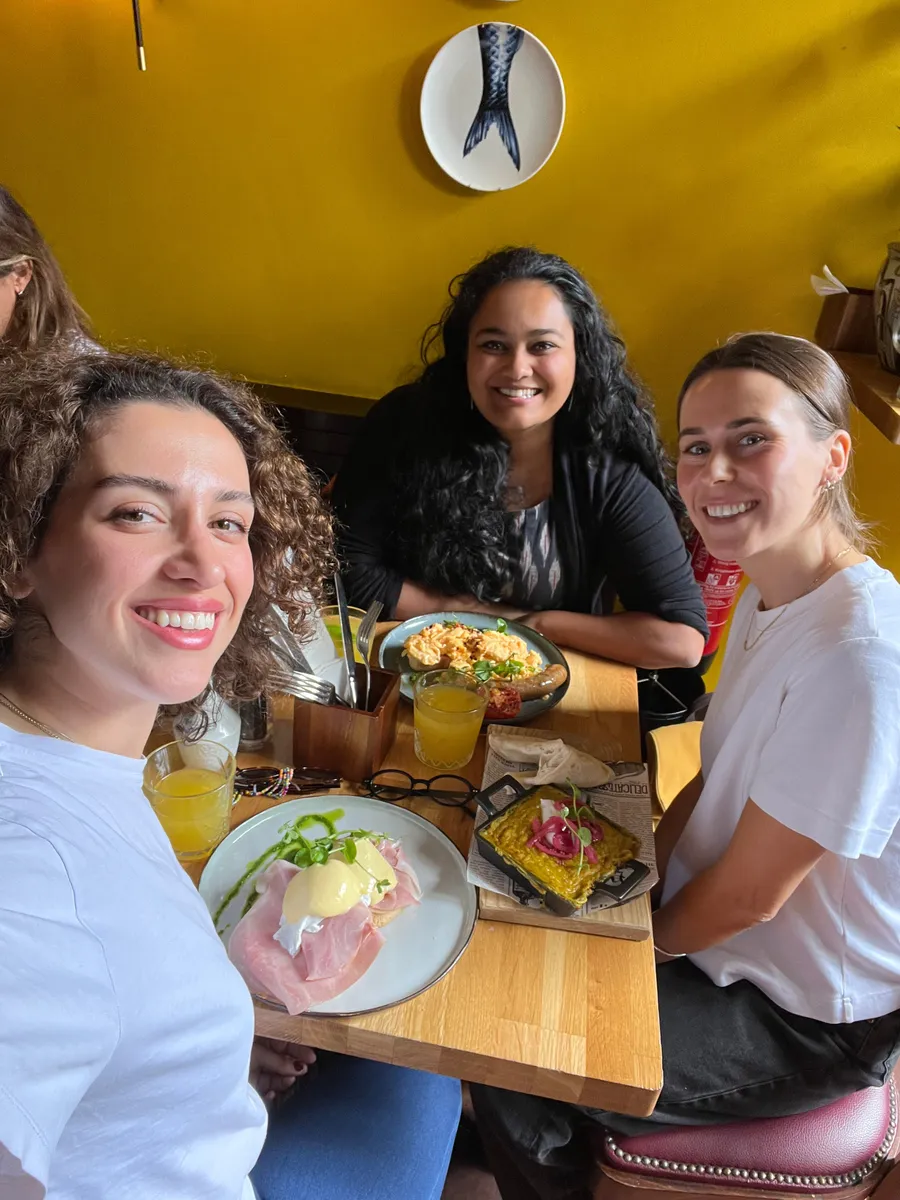 Four women smiling at a restaurant table with dishes and drinks, yellow wall background, mermaid tail decoration visible.
