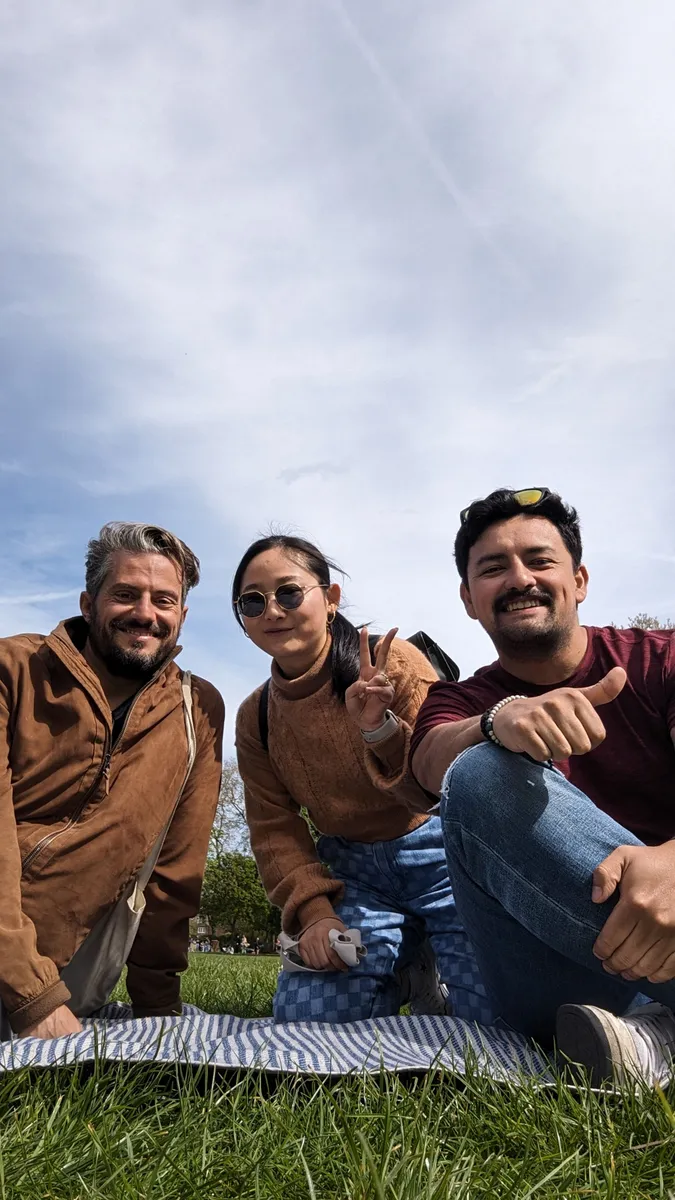 Three smiling people posing outdoors on grass with a picnic blanket, cloudy sky in background