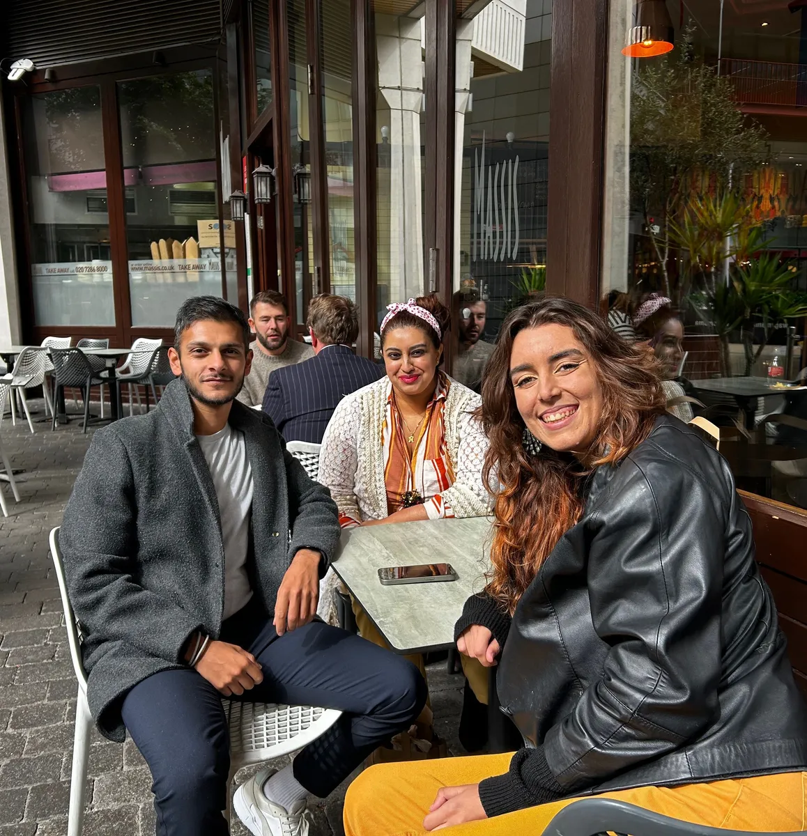 Three smiling people at an outdoor café table during a Creative Lunch Club meetup in London, with others visible in the background.