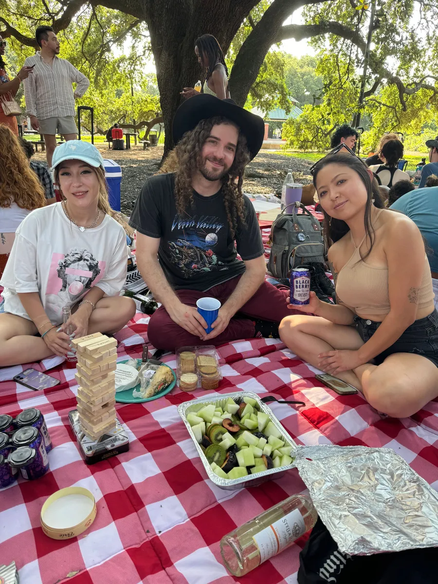 Three people enjoying a picnic lunch under a large tree in a Houston park with food and drinks on a red checkered blanket.