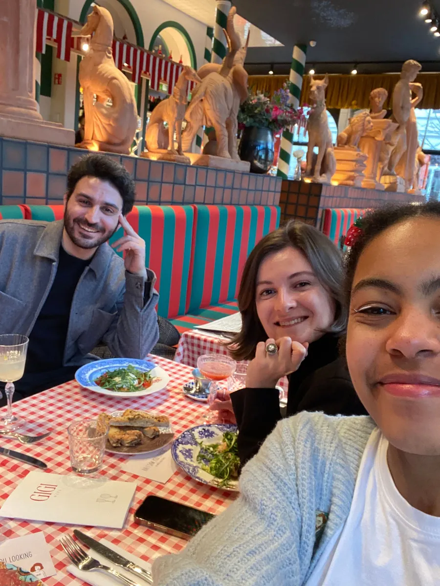 Three people smiling at a red and green striped booth during a Creative Lunch Club meetup in Ghent, with decorative statues and festive decor in background.