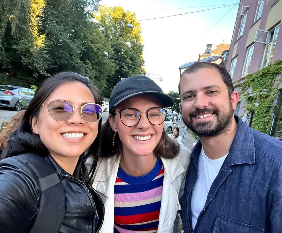 Three smiling people at a Creative Lunch Club meetup in Copenhagen, standing outdoors with buildings and trees in background.