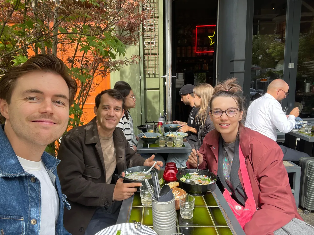 Group of creative professionals dining outdoors at a Berlin restaurant with neon signage and autumn foliage visible.
