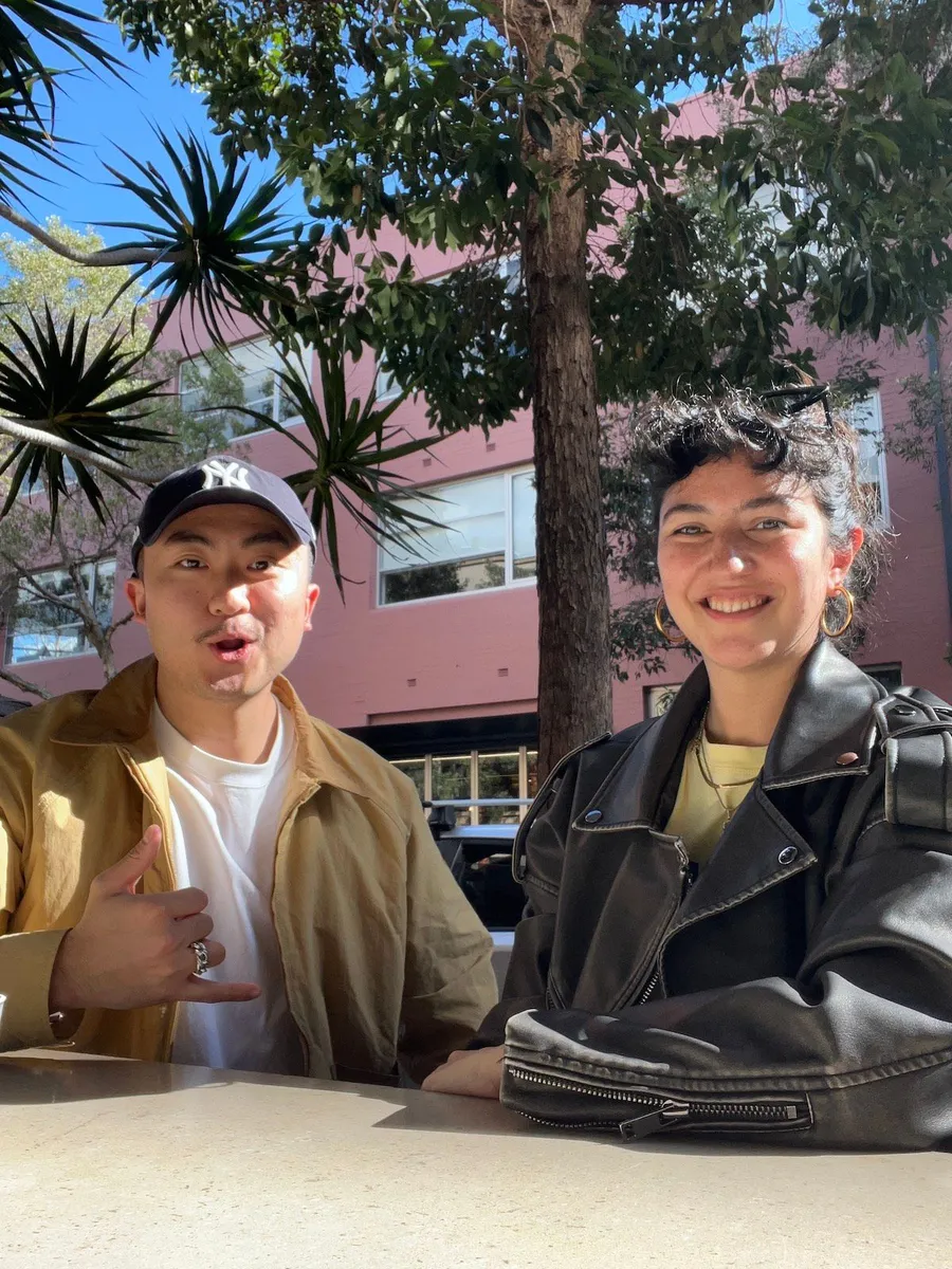 Two young people smiling at a Creative Lunch Club meetup in Sydney, seated outdoors with a pink building and palm trees in the background.