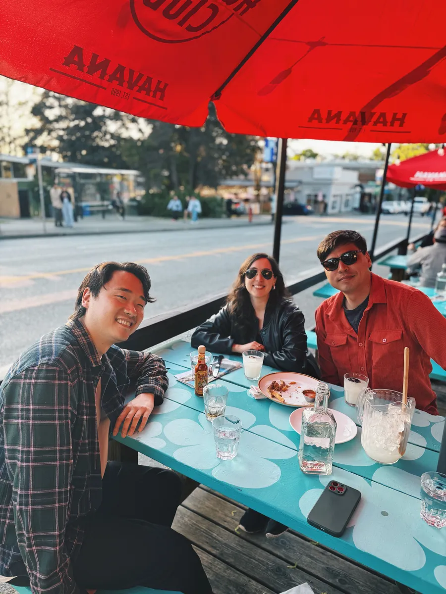 Three people dining outdoors under red umbrellas at a turquoise patterned table in Vancouver during daytime.