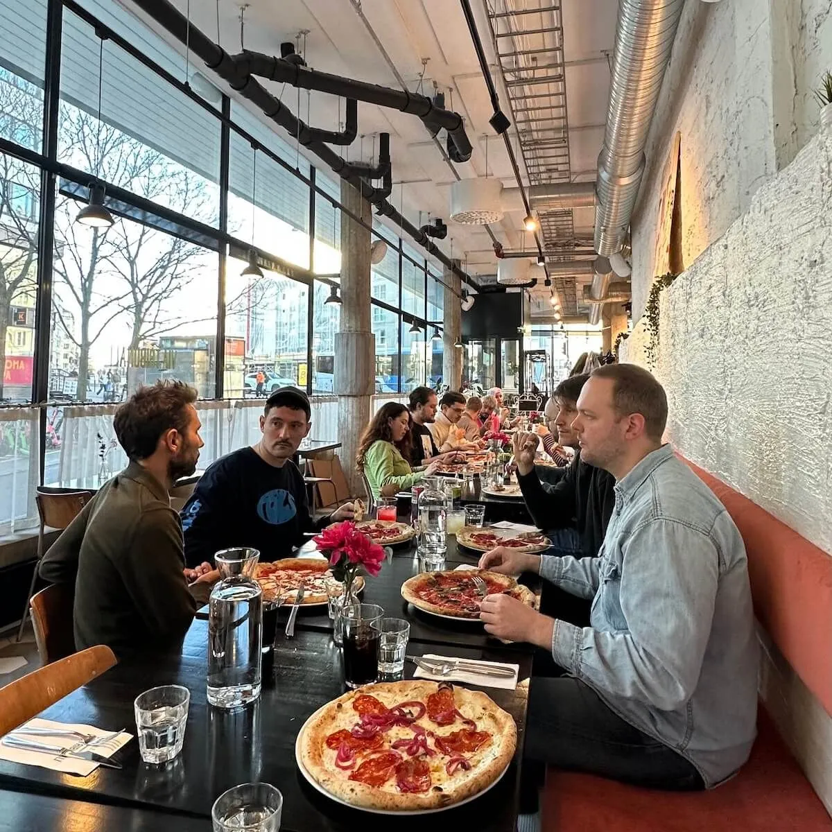 Group of professionals dining together at a modern pizzeria with large windows overlooking Helsinki's cityscape during daytime.